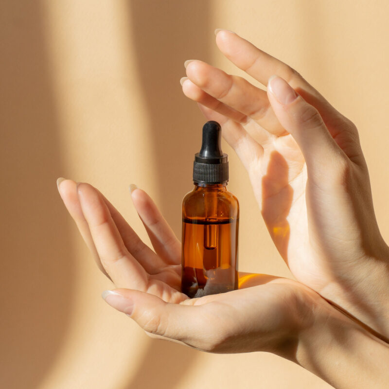 Women's Hands Hold Bottle Of Amber Glass With Cosmetic Serum On Brown Background
